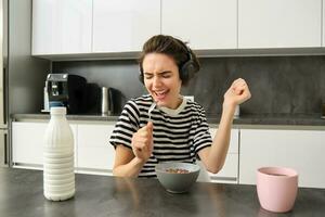 Happy and emotional young woman, singing while eating breakfast, having her cereals with milk, listening to music in wireless headphones, sitting in the kitchen photo