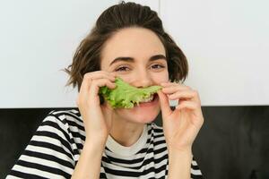 cerca arriba retrato de joven mujer, vegetariano chica, gustos comiendo verduras, posando con lechuga hoja y sonriente, posando en el cocina. concepto de sano comida y dieta foto