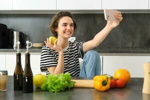 retrato de hermosa sonriente mujer en el cocina, tomando selfie con manzana, Cocinando sano comida desde verduras, el cortar comida ingredientes y grabación vídeo en móvil teléfono foto