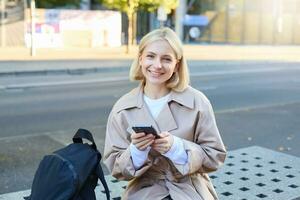 Portrait of smiling female model sitting on street bench with smartphone, waiting for friend, ordering a ride using mobile app photo