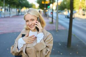 retrato de alegre, joven sincero mujer caminando en calle, respuestas teléfono llamar, negociaciones en teléfono inteligente y en pie cerca camino, sonriente y mirando contento foto