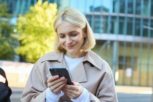 Close up portrait of smiling female model on street, sitting on bench with mobile phone, looking at smartphone screen, messaging with friend while waiting outside photo