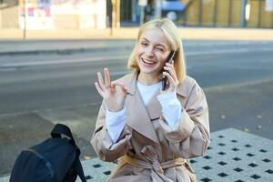 Image of young woman talking on mobile phone, sitting on bench outside, showing okay, ok approval gesture, made a deal over the telephone photo