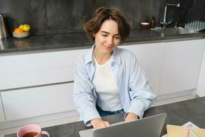 Young woman works from home, uses laptop in her kitchen. Student doing homework. Girl studies, watches online webinar, does distance learning photo