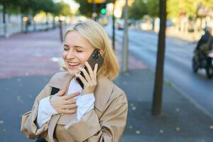 cerca arriba retrato de hermosa joven mujer, rubio niña caminando en calle con móvil teléfono, chateando con amigo, tiene contento cara expresión mientras hablando terminado Teléfono móvil en su camino foto