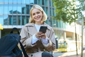 Lifestyle portrait of cute blonde girl with smartphone, sitting on bench with backpack, using mobile phone, reading message photo