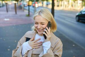 Close up portrait of smiling, happy and carefree woman on streets of big city, laughing while talking on mobile phone, chatting on smartphone and walking photo