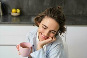 contento mañanas. retrato de contento morena mujer, bebidas taza de café en su cocina y sonriente, acogedor y calentar comienzo de el día con taza foto