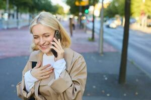 Close up portrait of happy girl laughing, standing on street with smartphone, talking on mobile phone, giggle and smiling photo