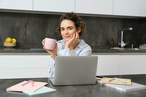 Self-employed woman works from homes, sits with laptop and drinks coffee in kitchen, surrounded with documents. Girl student studies remote, does project on computer, holds a cup photo