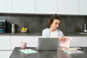 Portrait of working woman at home, checks her notebook, flips page, connects to online meeting via laptop. Girl learns programming remotely, studies course in internet photo