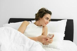 Portrait of young woman waking up in bed, using smartphone, turn off alarm on mobile phone, lying in her bedroom on white linen sheets photo