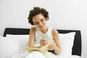 Image of mindful young woman sitting in bed, waking up and writing in journal, making notes, sharing her thoughts in diary photo