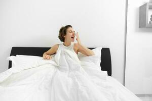 Image of smiling, beautiful modern woman wake up in her bed, yawning with pleased face, had a good night sleep photo