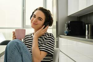 Portrait of tender, beautiful woman with tea, listens music in headphones, holds cup and smiles photo