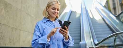 Portrait of young stylish woman, female employee, standing near escalator, using mobile phone, sending message on smartphone, tap screen, smiling and reading on telephone photo