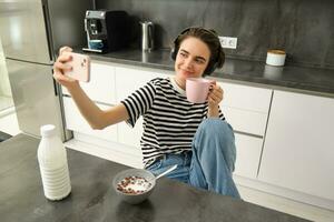 Portrait of modern happy woman, taking selfie, lifestyle blogger recording video of her eating breakfast and drinking tea in the kitchen, using smartphone for making content photo