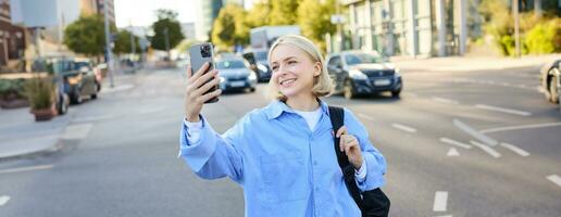 Image of smiling young woman, modern girl with smartphone, taking selfie, shooting a picture of busy city streets, recording video outdoors photo