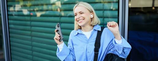 Carefree woman with backpack and mobile phone, wearing wireless earphones, listening to music and smiling, standing on street in city centre, laughing photo