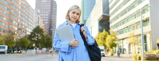 Portrait of young confident woman, college student with backpack and laptop, heading to lesson, standing outdoors on empty street with big buildings behind photo