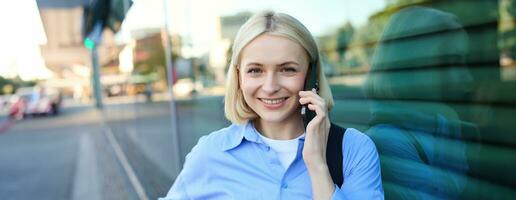 Close up portrait of smiling blond woman, chatting on the phone, talking on mobile telephone, standing on street outdoors photo