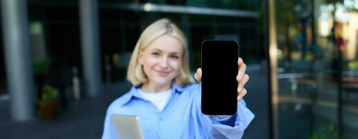 Portrait of beautiful young woman, student posing on street near campus, holding laptop, showing mobile phone screen, demonstrating app or promo on smartphone and smiling photo