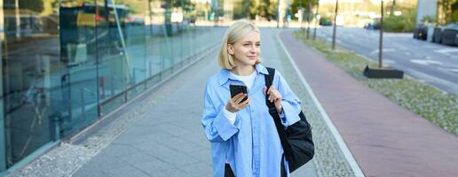 Portrait of blond young woman, using navigation map app on smartphone, walking along street, using directions on phone, holding backpack photo
