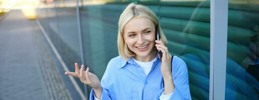 cerca arriba retrato de sonriente rubio mujer, chateando en el teléfono, hablando en móvil teléfono, en pie en calle al aire libre foto