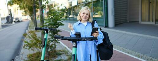 Beautiful blond girl, scans QR code on mobile phone, stands near electric street scooter to rent it, requesting a ride, smiling and looking happy photo