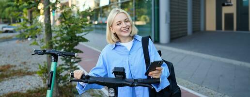 Portrait of smiling blond woman, college student using smartphone app to scan QR code on green electric scooter on street, renting a ride photo