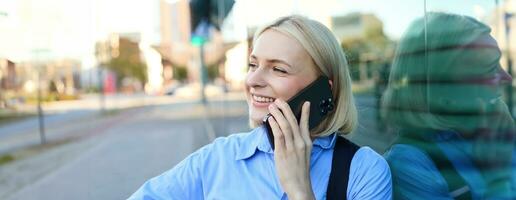 Lifestyle portrait of happy blond woman, chatting over the phone, answers a call, laughing and smiling, standing on street and looking cheerful photo