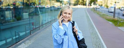 Portrait of young smiling woman talking on mobile phone, student walks home with smartphone, carries black backpack photo