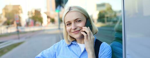 Close up portrait of smiling blond woman, chatting on the phone, talking on mobile telephone, standing on street outdoors photo