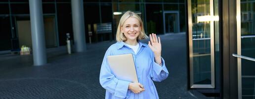 Friendly smiling student, blond girl with notebooks, tutor waving hand at camera, saying hello, standing near office building outdoors photo