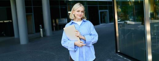 Stylish, modern young woman, student standing on street in campus, heading to lecture with notebook and study material, smiling at camera photo