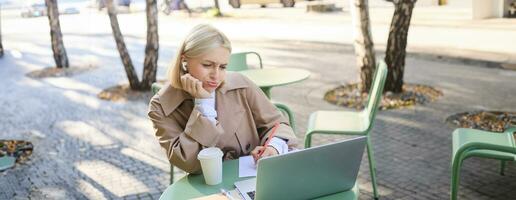 Portrait of blond girl with upset face, listening lecture, attends online course in wireless headphones, sits in outdoor cafe and frowns while makes notes, studying remotely, using laptop photo