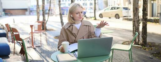 Image of young woman with puzzled face, looking at laptop in wireless headphones, confused while listening to lecture or online speaker, attend web course while sitting in outdoor cafe photo