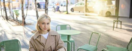 Vertical shot of young blond woman in wireless headphones, making notes, studying online, attend web lecture, using laptop, writing in notebook, looking serious at camera photo