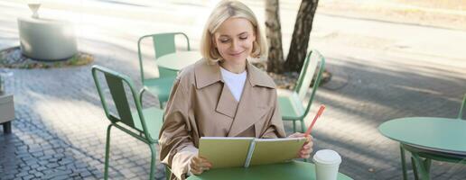 Portrait of young creative woman, doing sketches outdoors, sitting in coffee shop with notebook and pen, drawing art and smiling photo