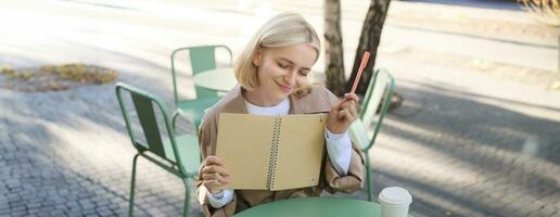 Portrait of young creative woman, doing sketches outdoors, sitting in coffee shop with notebook and pen, drawing art and smiling photo