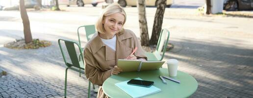 Image of young smiling woman, sitting in outdoor cafe, doing sketches, drawing art on street, spending time on coffee shop and creating art photo