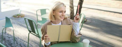 Portrait of young beautiful blond woman, artsy girl in coffee shop, holding notebook and pen, writing in her journal, drawing sketches outdoors photo