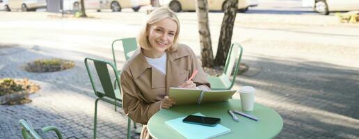 Portrait of beautiful blond woman, sitting in outdoor coffee shop, drawing in cafe in notebook, making sketches outside on street photo