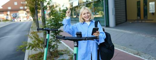 Portrait of young smiling female model, renting an electric scooter, using mobile phone to scan QR code with smartphone app, riding home from university, standing outdoors on street photo