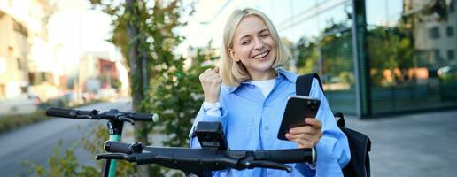 Portrait of smiling blond woman, college student using smartphone app to scan QR code on green electric scooter on street, renting a ride photo