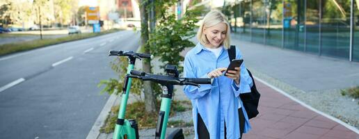 Image of smiling young woman heading to work, renting, using mobile phone app to unlock street scooter to get to her destination, posing on street with backpack photo