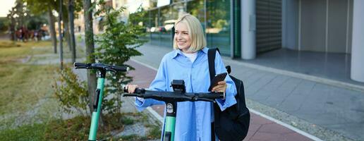 Portrait of young woman, renting a scooter, using mobile phone app to unlock it, using quick ride to get to work, smiling and looking happy photo