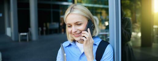 Happy female model, blond woman talking on mobile phone, chatting with someone, standing near office building with laptop and smartphone, smiling photo