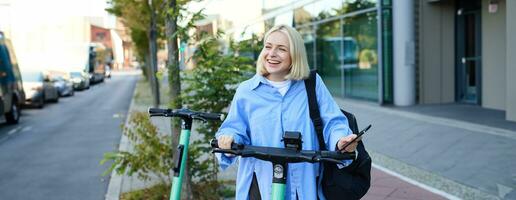 Portrait of smiling blond woman, college student using smartphone app to scan QR code on green electric scooter on street, renting a ride photo