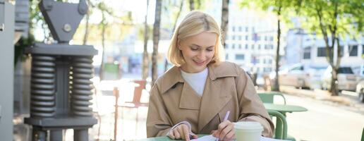 retrato de sonriente hermosa mujer, escritura en su diario o computadora portátil, haciendo deberes mientras sentado en café y Bebiendo café foto